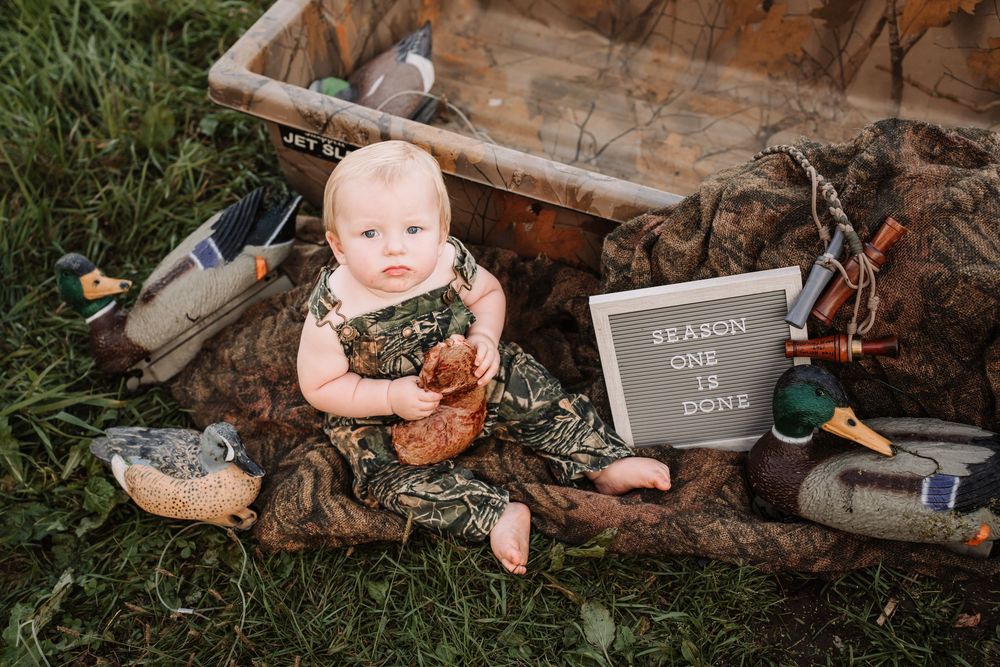 A rustic hunting-themed cake smash photo session with decoy ducks and vintage props in an outdoor woodland setting.