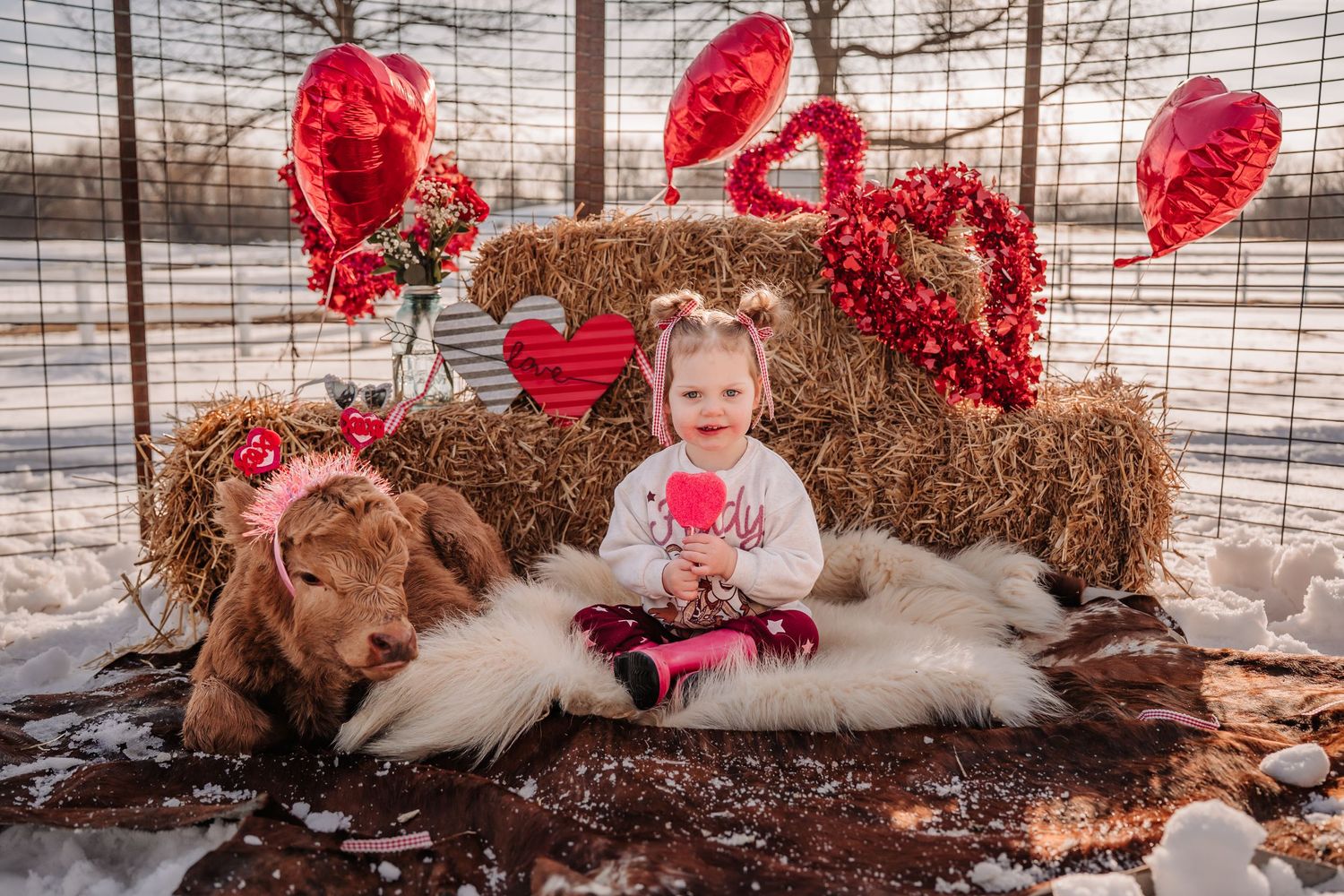 Toddler girl sits on fur rug beside hay bales with Valentine hearts and red balloons in snowy outdoor setting.
