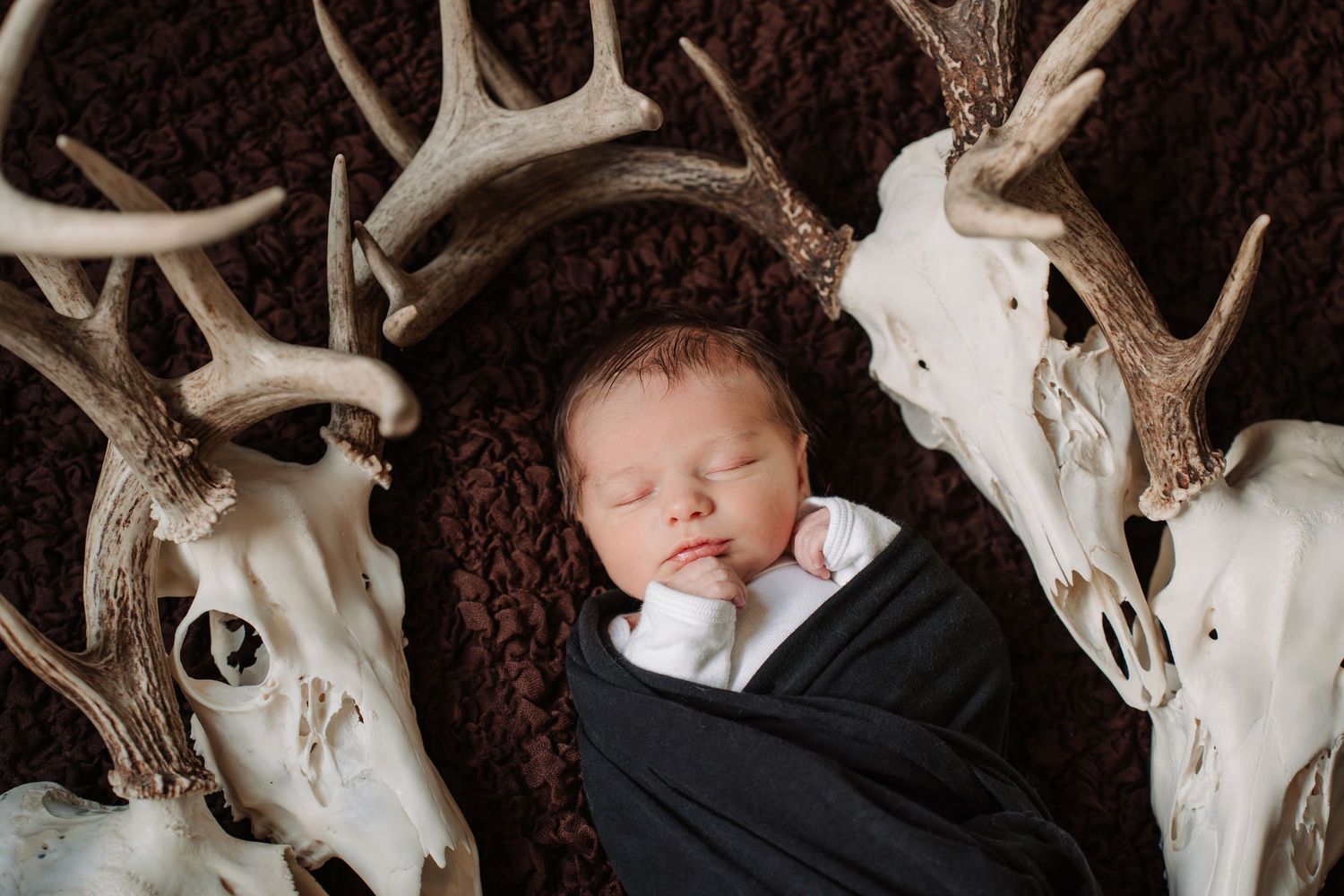 Newborn baby swaddled in black wrap sleeping peacefully among decorative deer antlers and skulls on brown fur backdrop.