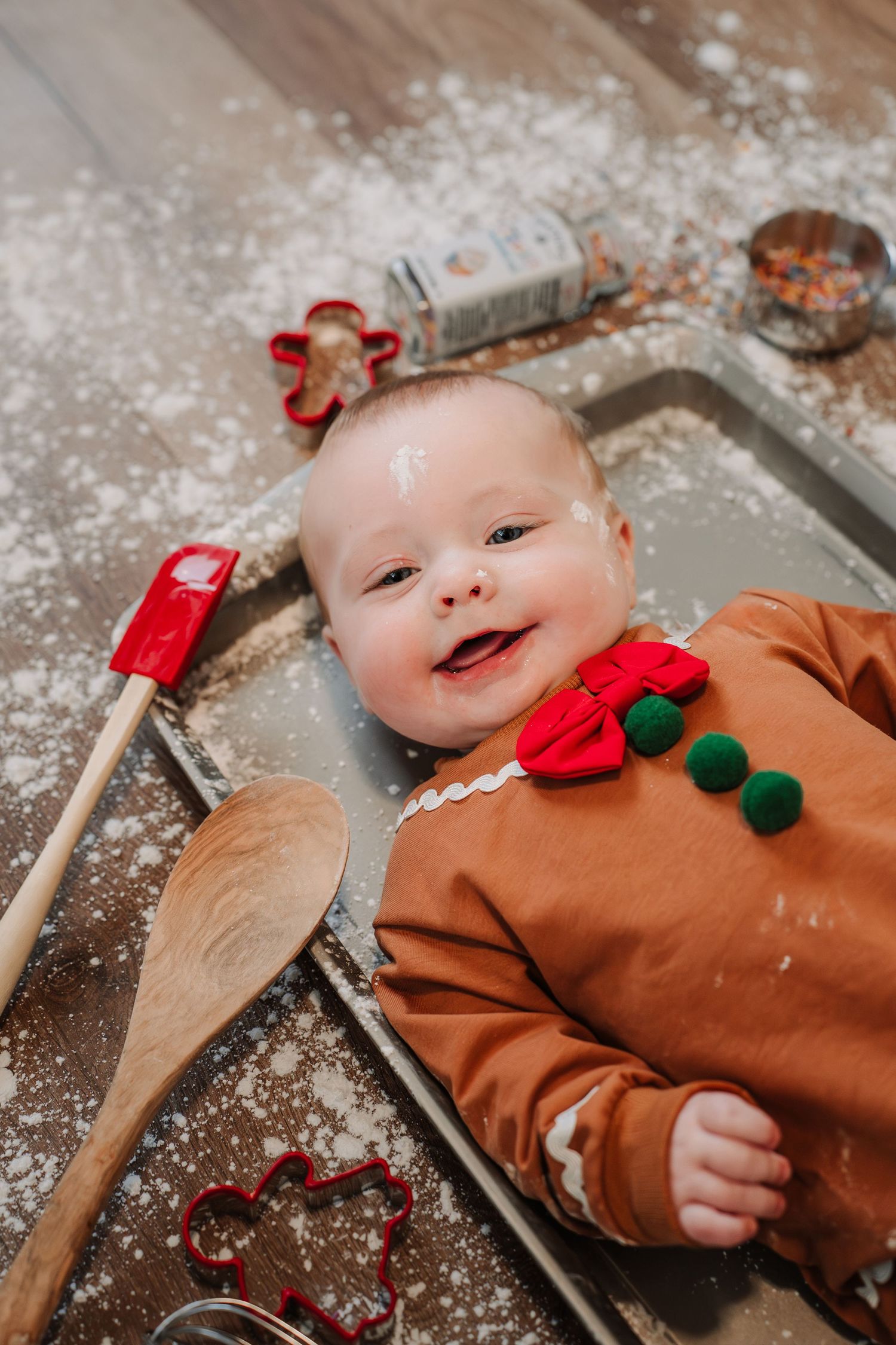 Baby in brown outfit lying on flour-dusted surface with holiday decorations and baking utensils.