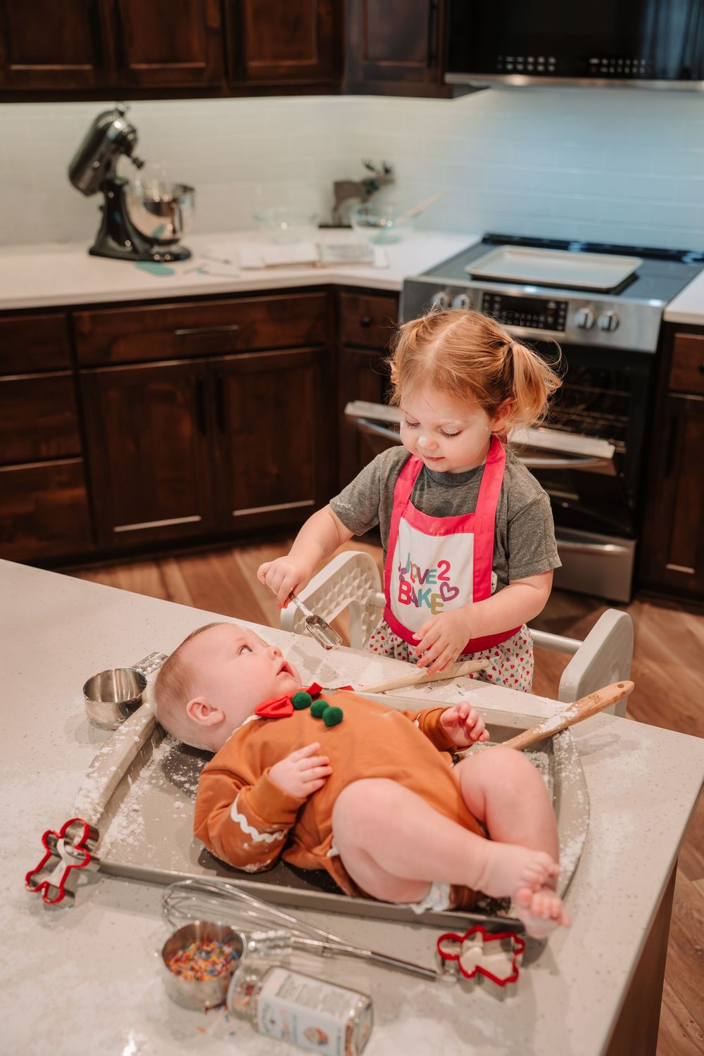A child in a red apron decorates a gingerbread baby cookie on a kitchen counter during holiday baking.