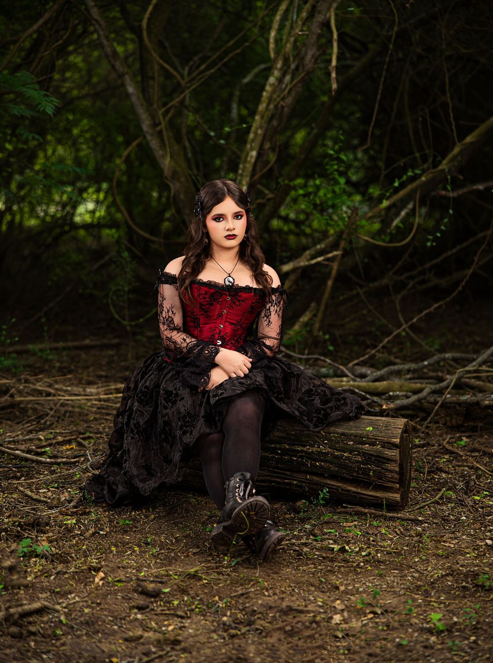 Young woman in red corset and black gothic dress sits on a log in a dark forest setting.