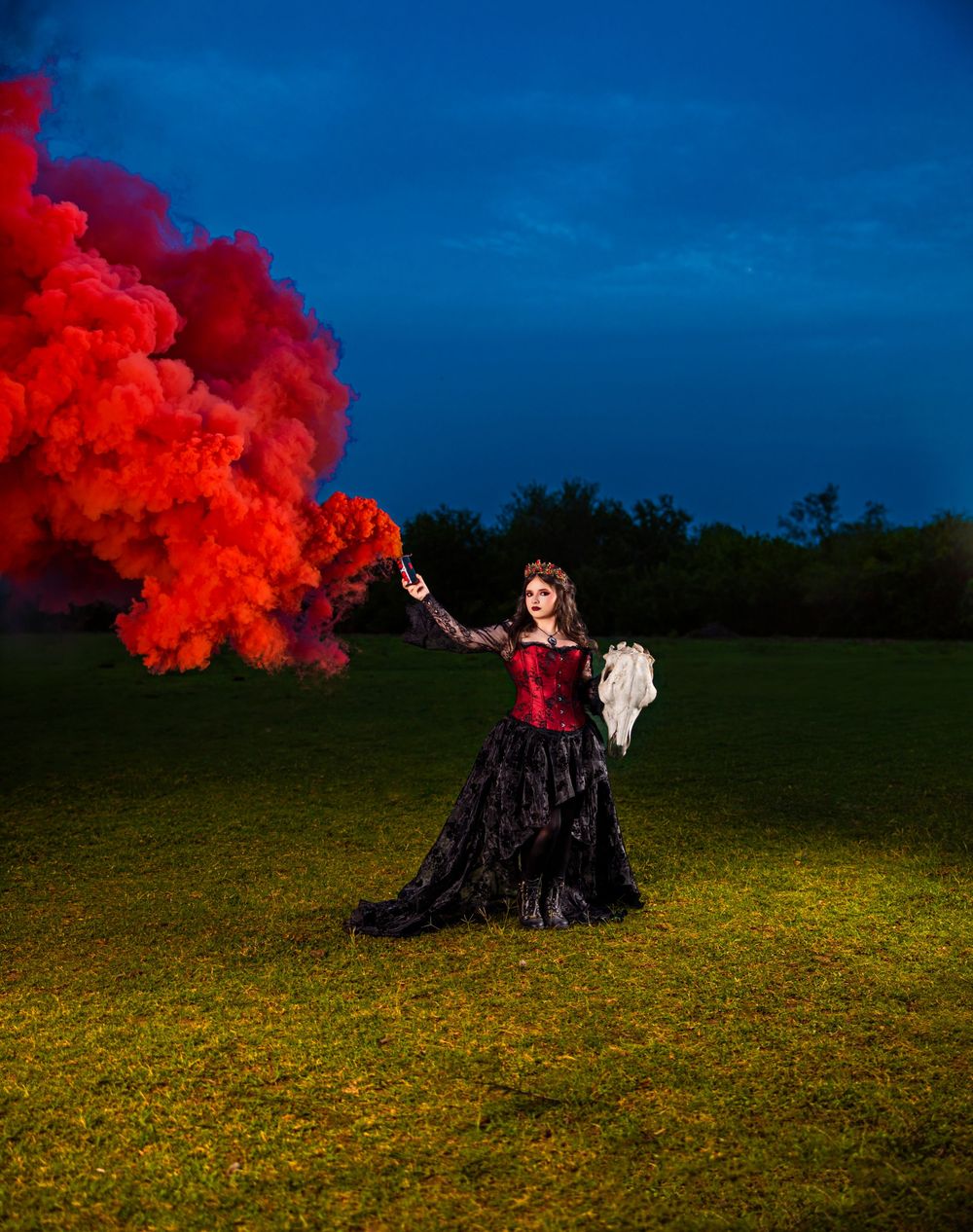 Woman in black gothic gown holds red smoke flare dramatically in green field under a deep blue evening sky.