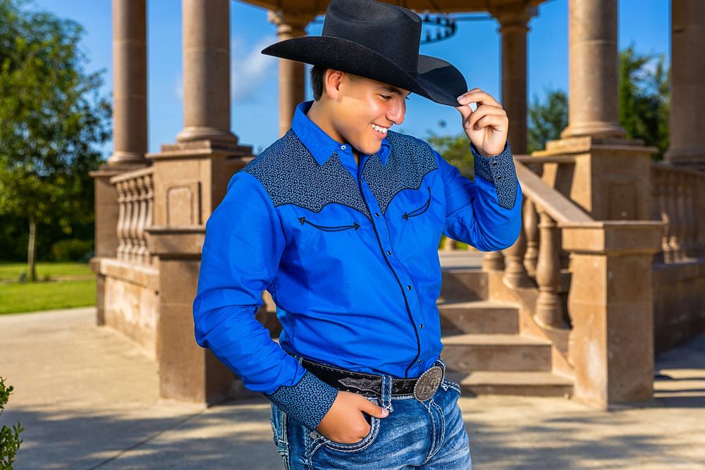Person in bright blue western shirt and black cowboy hat poses against stone pillars in outdoor setting.