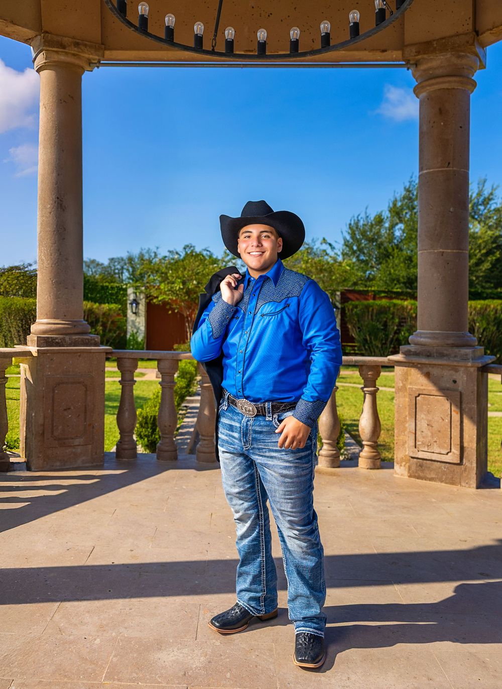 A person wearing a cowboy hat and blue denim stands in front of a stone gazebo with columns in a sunny outdoor setting.