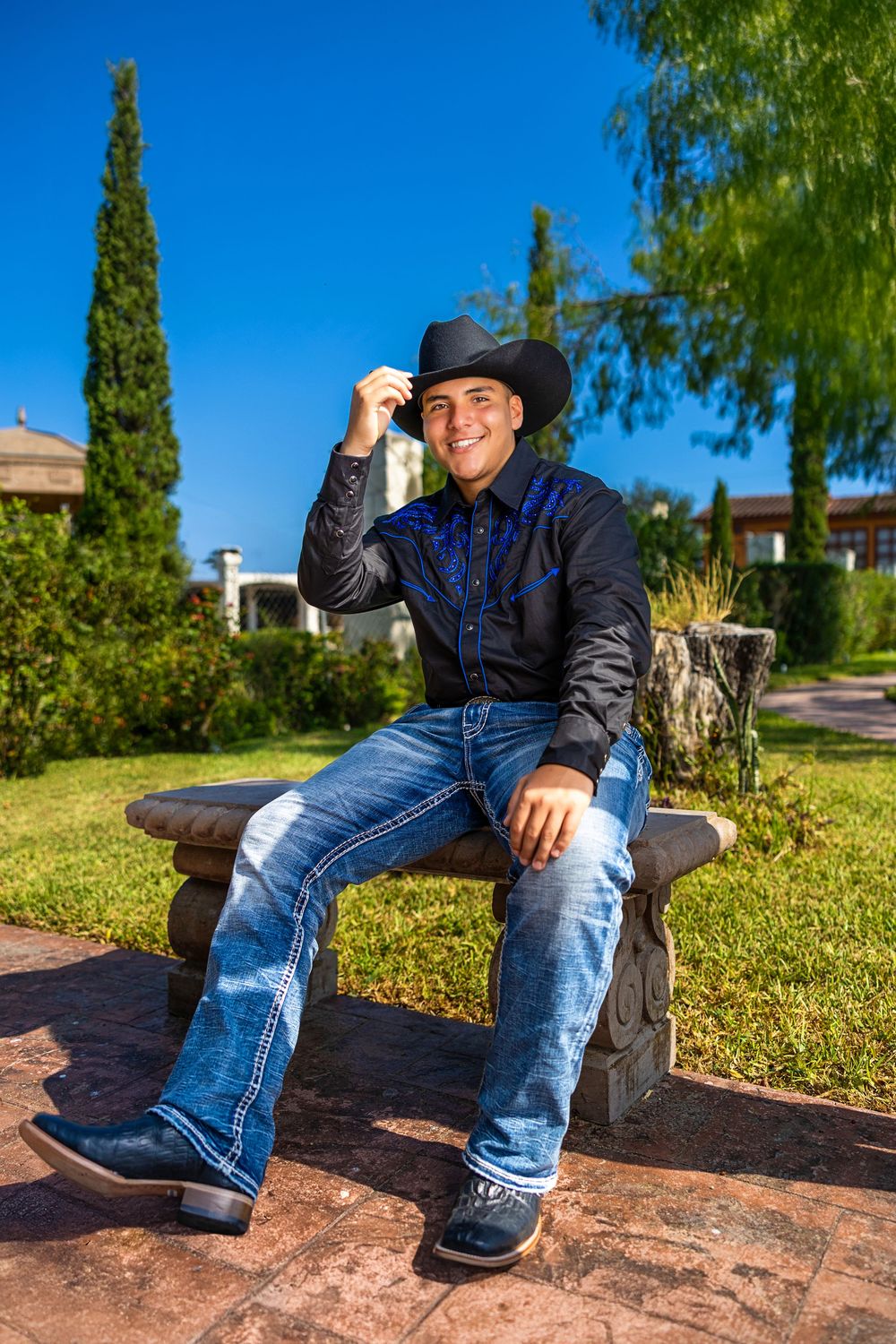 A person in blue jeans and boots sits casually on a stone wall with lush green trees in the background.