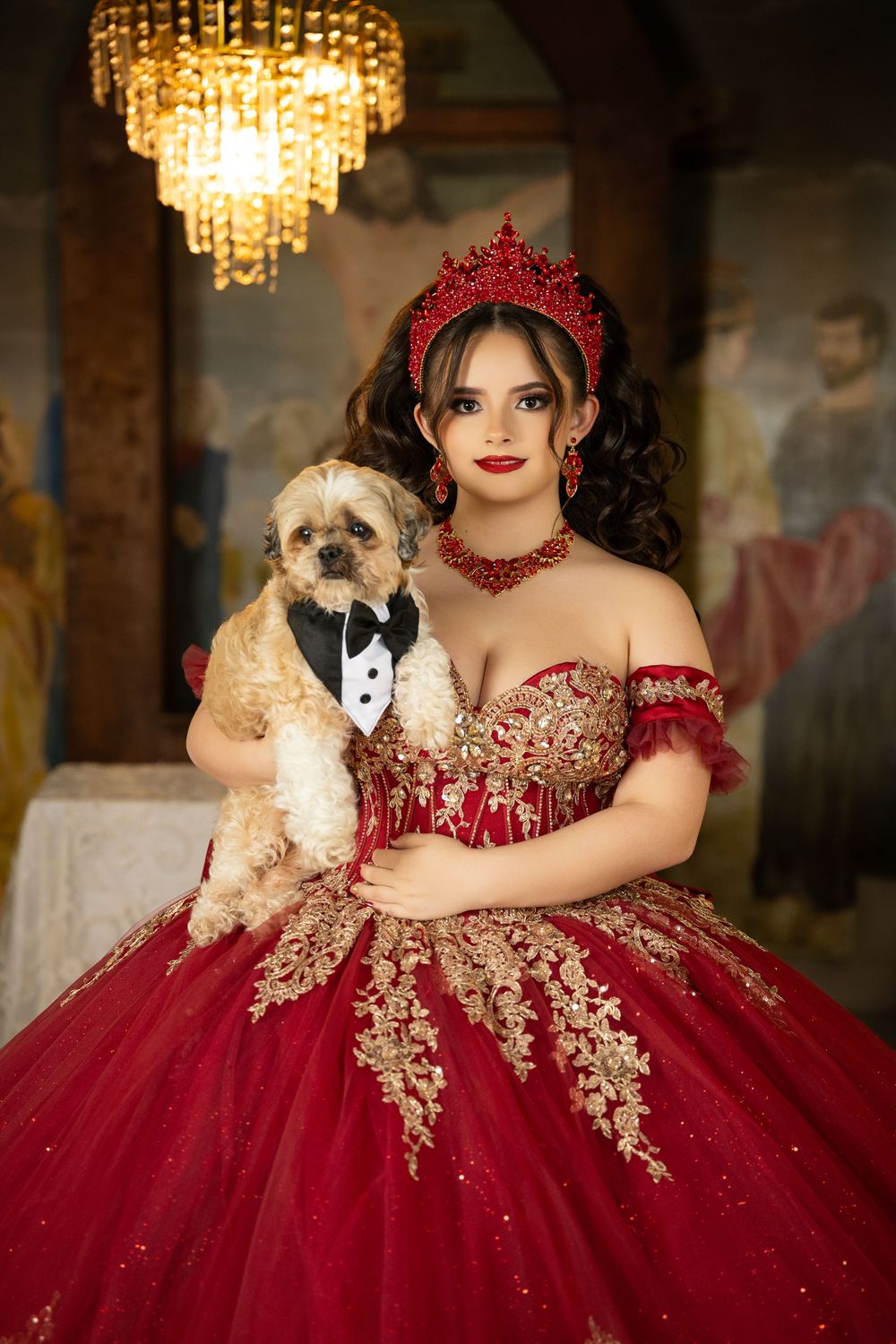 Young woman in red and gold quinceañera gown holding a small dog in a tuxedo, wearing a red crown and jewelry.