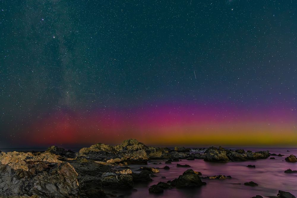 Rocky coastline under a stunning night sky with vibrant aurora borealis in pink, red, and yellow hues and Milky Way stars.
