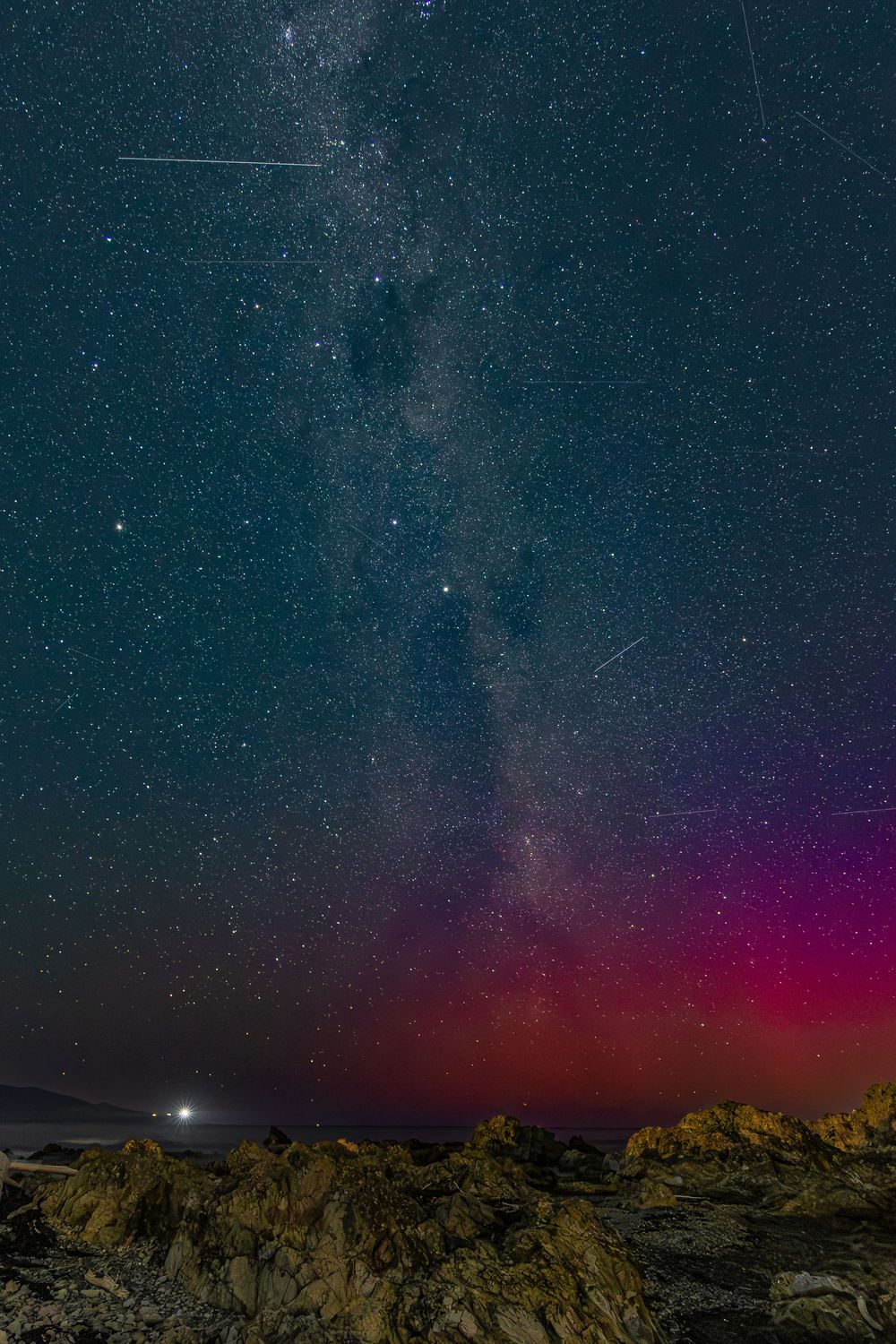 Milky Way galaxy over rocky coastline with vibrant pink aurora borealis and shooting stars in a stunning night sky.
