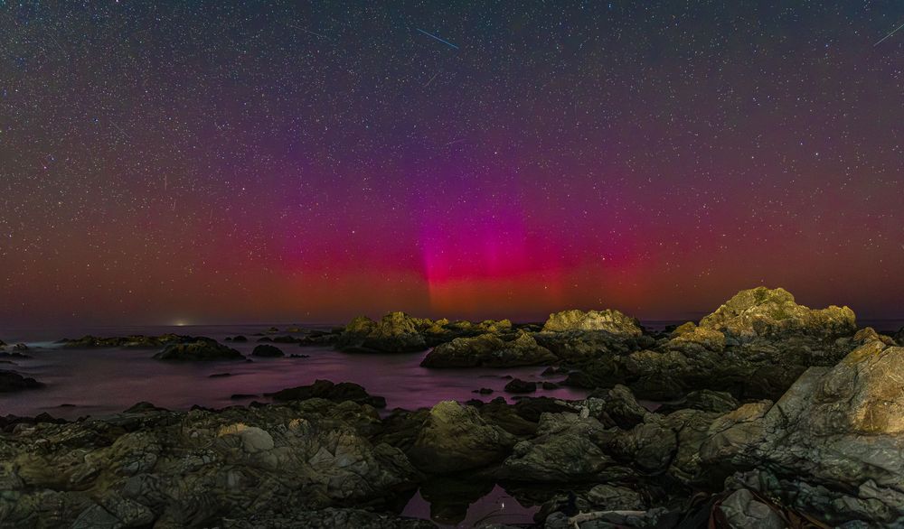 Aurora borealis illuminates rocky coastline with vibrant pink and red hues against a star-filled night sky.