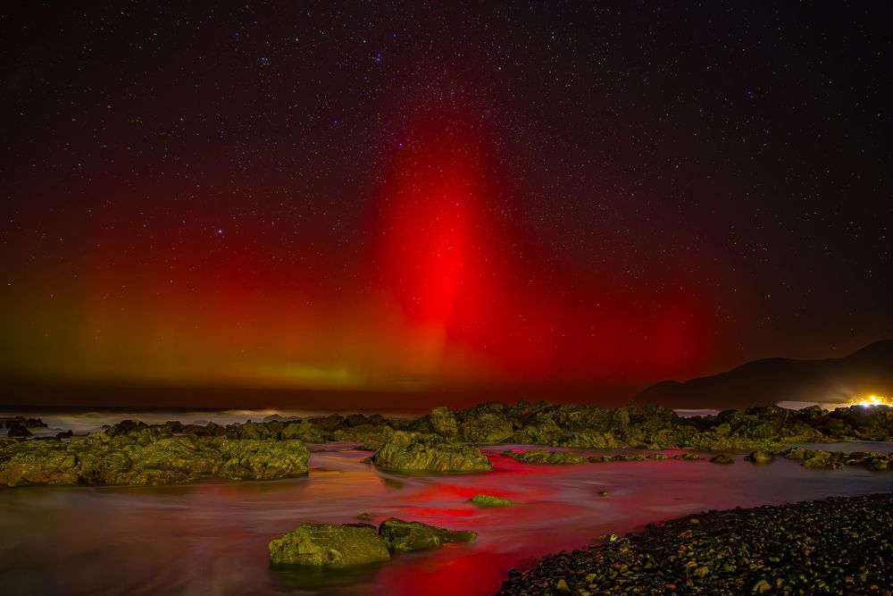 Vibrant red aurora borealis illuminates the night sky over a rocky coastal landscape with mossy rocks and reflective water.