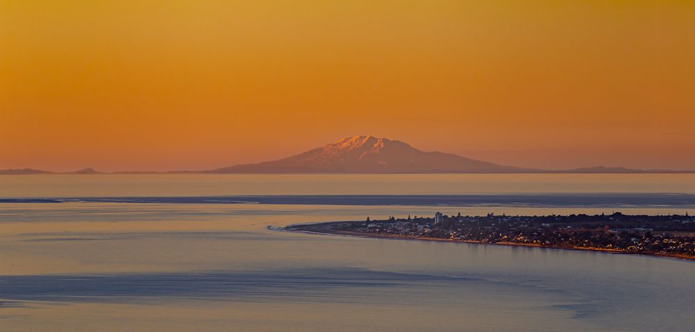 Vibrant orange and purple sunset sky reflects over calm ocean waters with distant mountain silhouettes on the horizon.