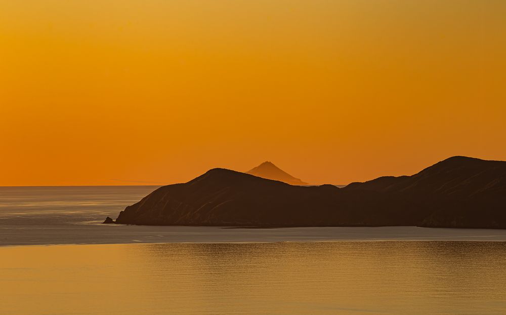 Dramatic orange sunset silhouettes mountain peaks against a glowing sky over calm coastal waters.