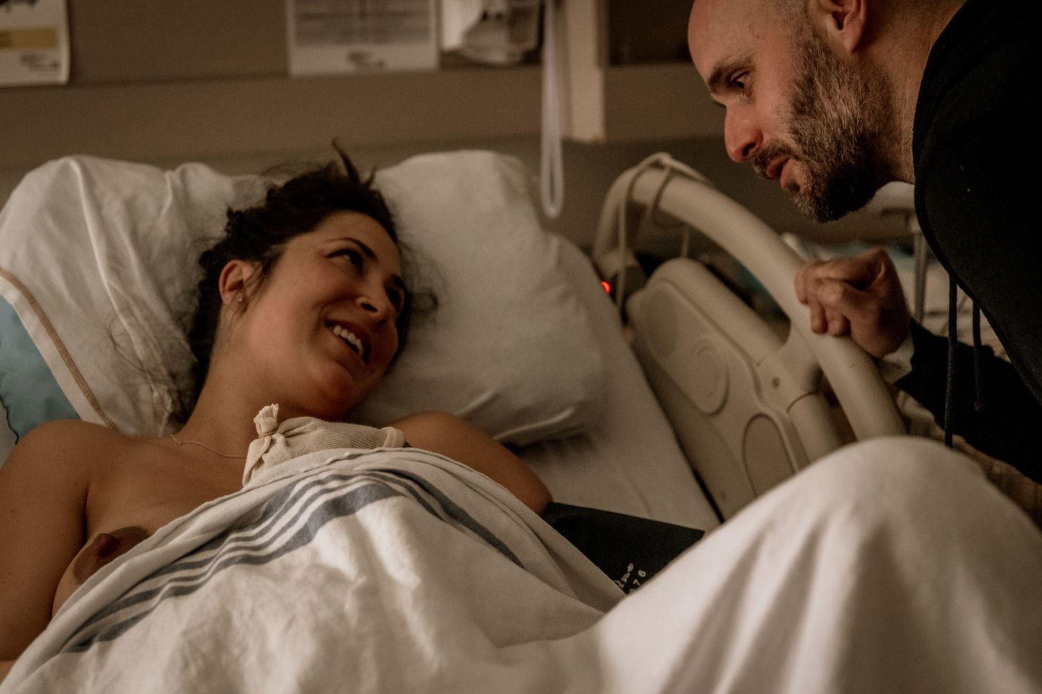 Couple shares an intimate moment in a hospital room as patient rests in bed with white linens.