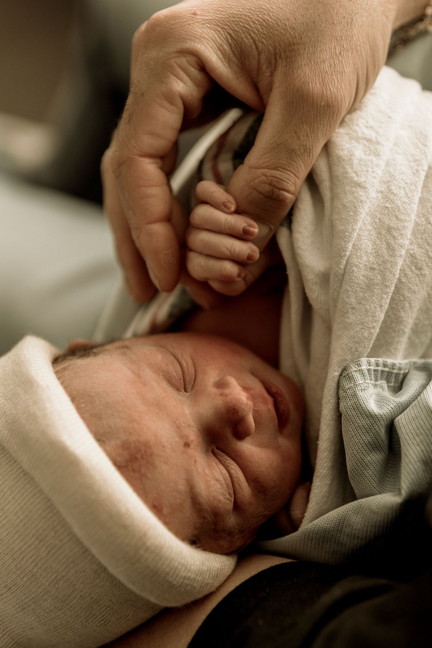 A newborn baby sleeps peacefully in white swaddling while a gentle hand tenderly touches their head.