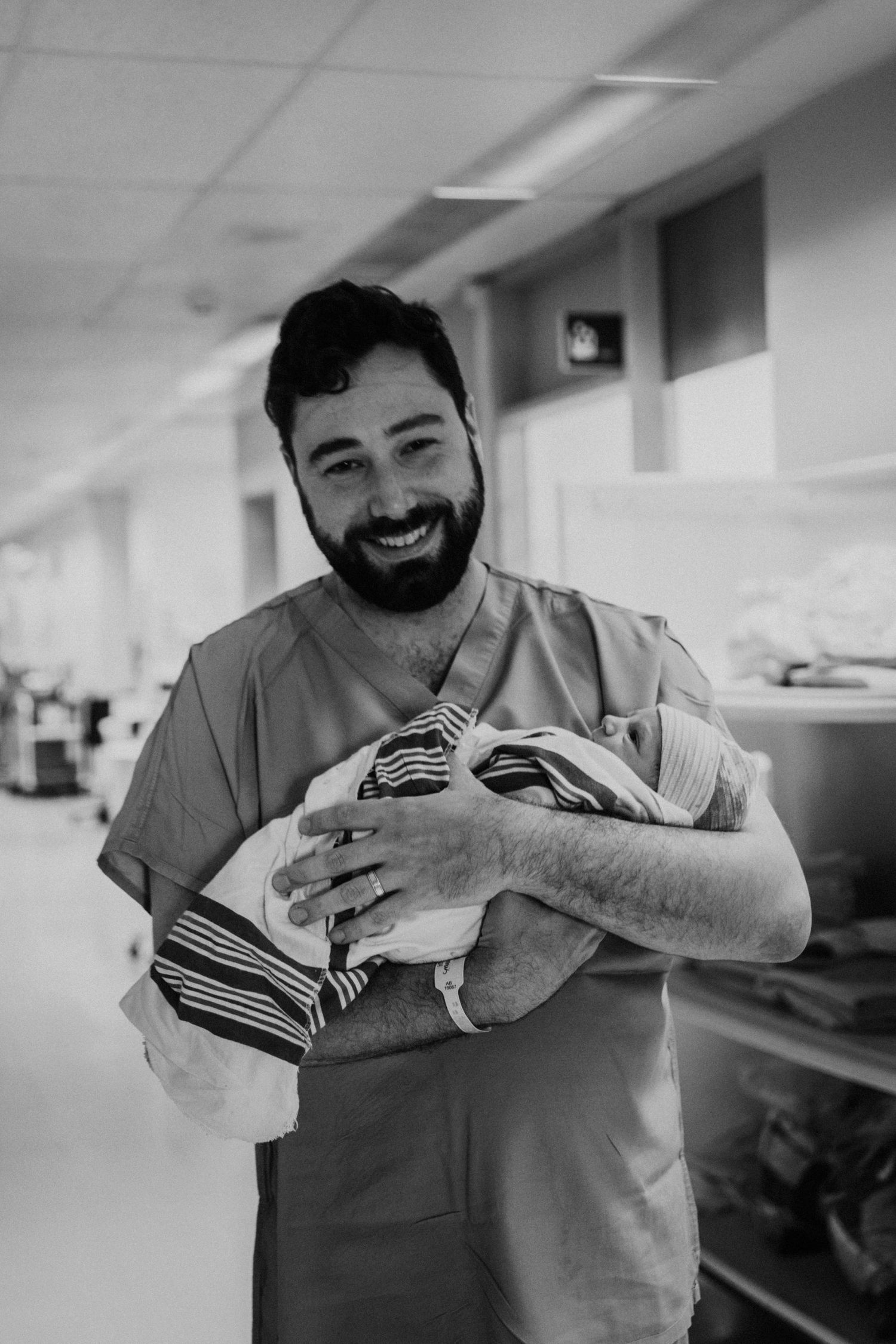 A healthcare worker in scrubs smiles warmly while holding a swaddled newborn baby in a hospital hallway.