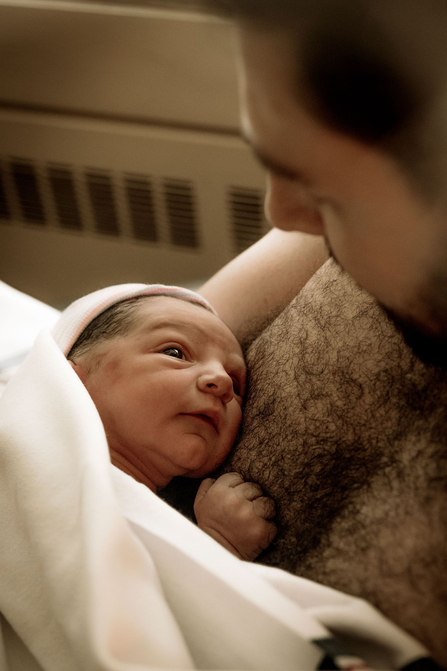 A newborn baby wrapped in white blanket gazes up while being held close.