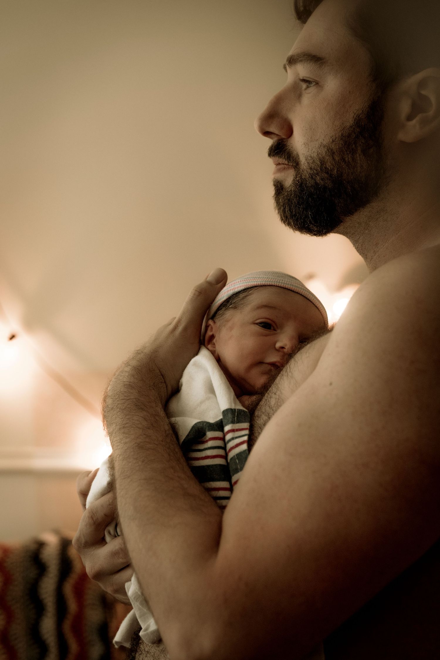 A tender intimate moment between a bearded person holding a sleeping newborn baby in soft warm lighting.