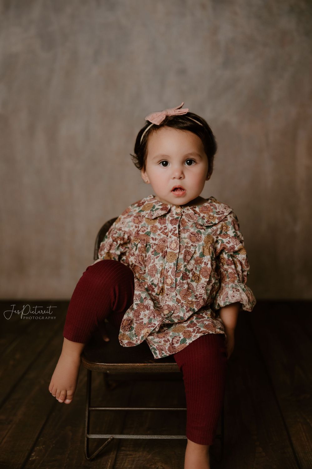 Portrait of toddler in floral top and burgundy leggings sitting on wooden stool.