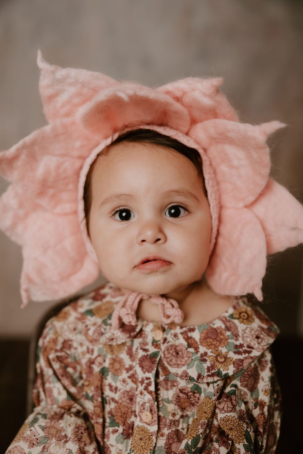 Baby wearing pink flower bonnet and floral dress against neutral backdrop.