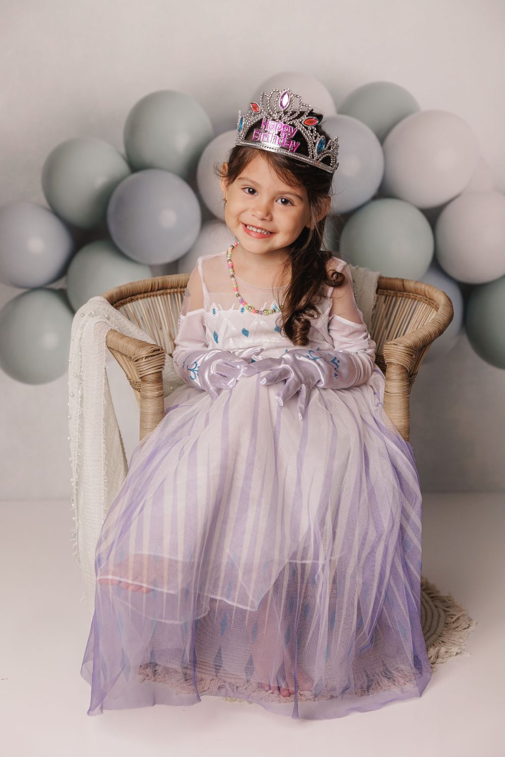 Elegant portrait in a wicker chair with silver balloon backdrop and flowing lavender princess dress.