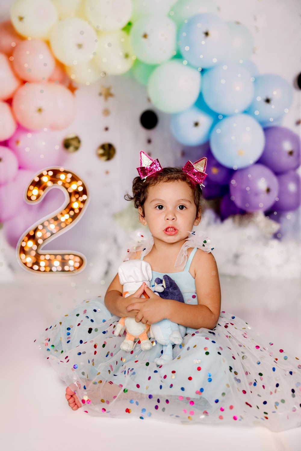 Birthday photoshoot setup with rainbow balloons, glowing number 2, and a polka dot tulle dress against a white backdrop.