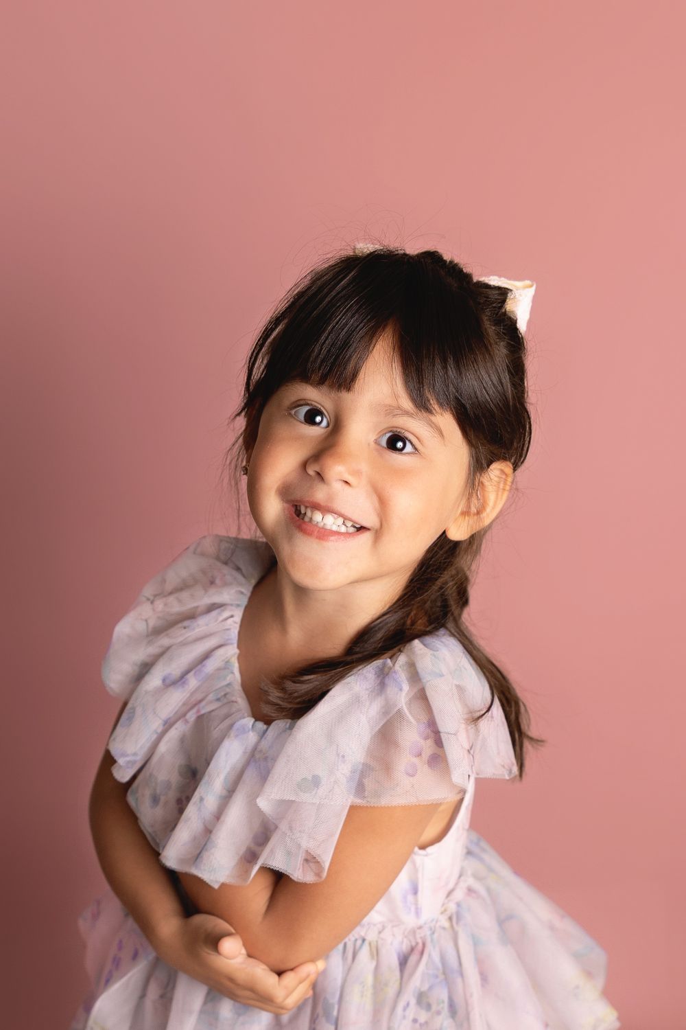 A young child wearing a light pink ruffled dress poses playfully against a pink background.