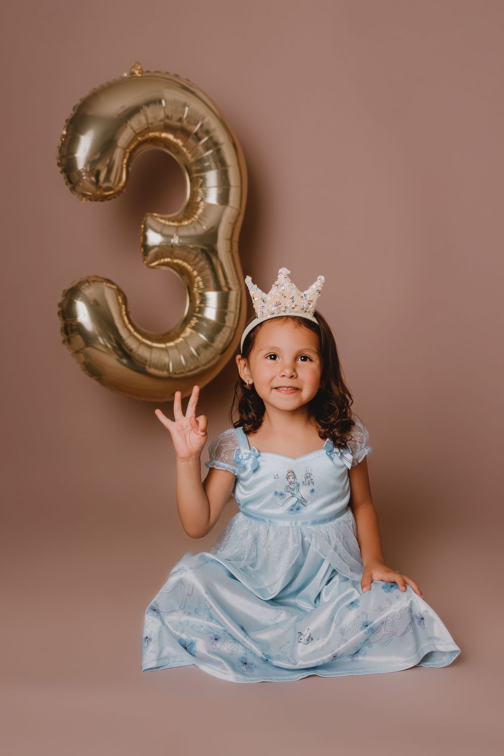 A child in a light blue princess dress poses with gold balloon number 3 against a pink backdrop for birthday photos.