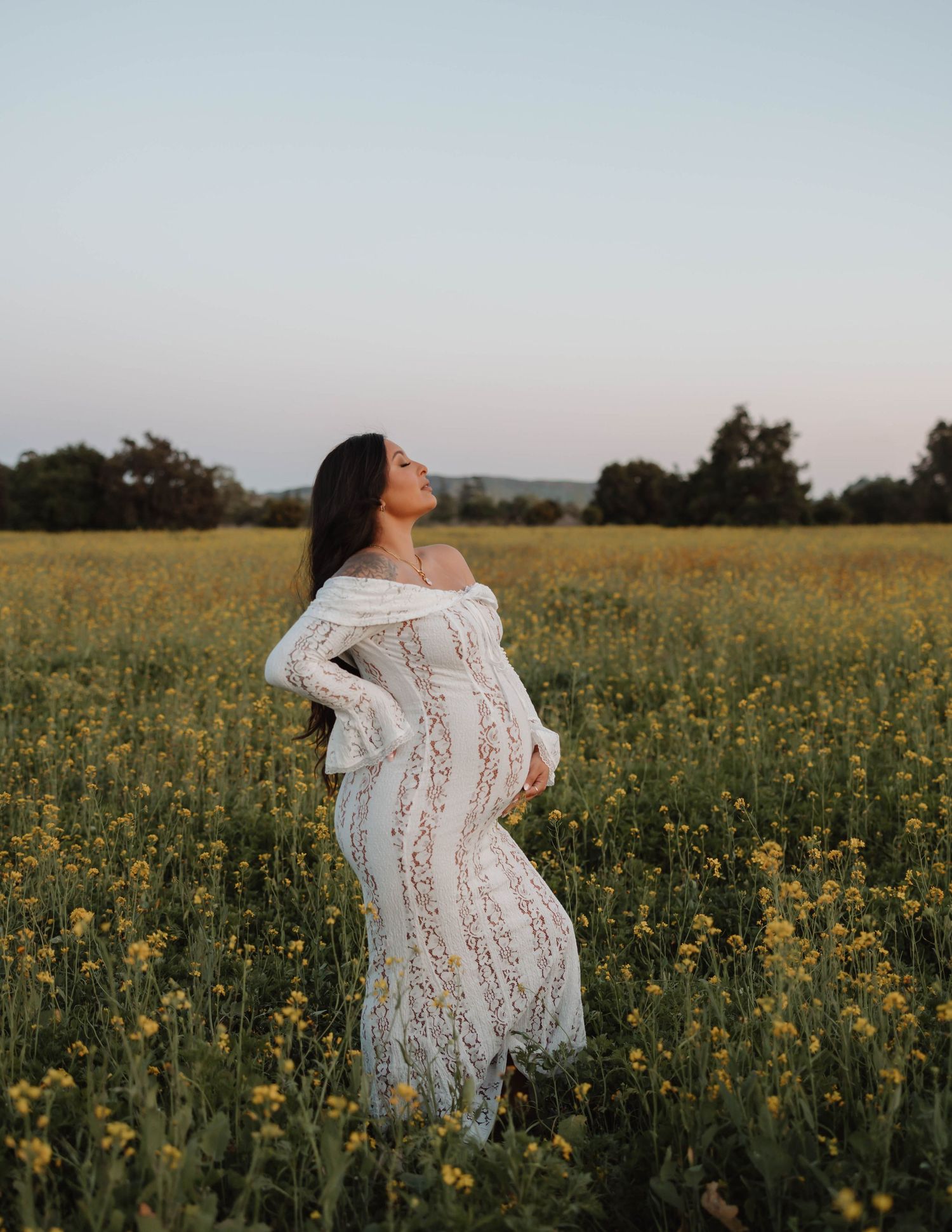 A woman in a white lace dress stands among yellow wildflowers during sunset for maternity photos in Orange County