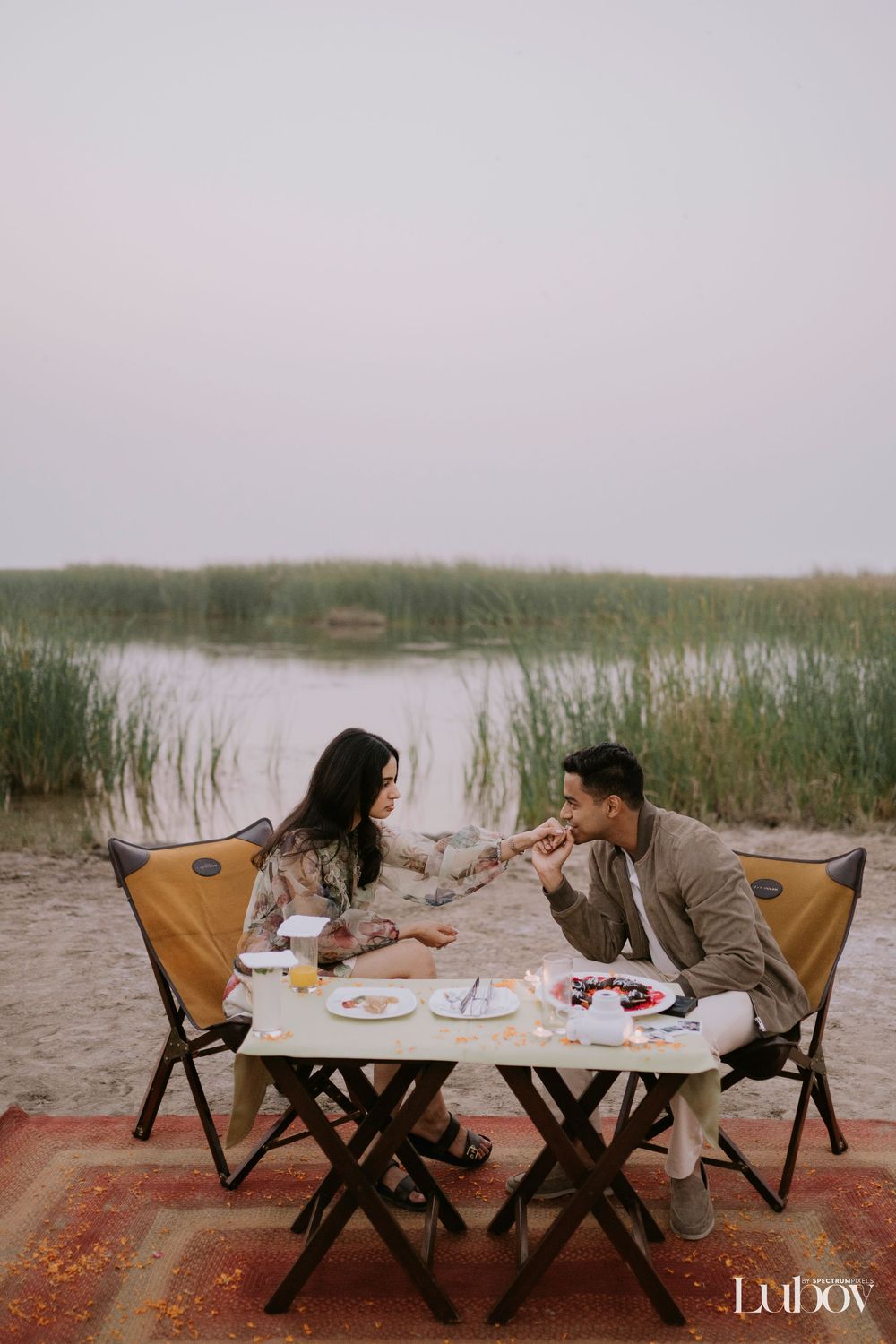 A couple enjoys a romantic lakeside picnic with camping chairs and a folding table at sunset.