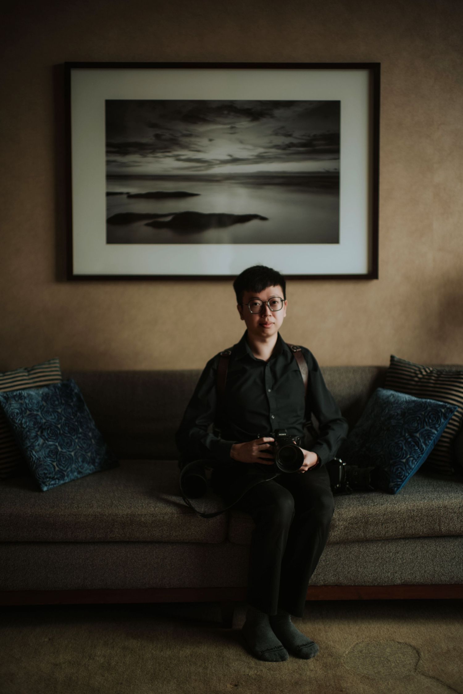 A photographer in casual attire sits on a couch beneath a black and white landscape photograph.