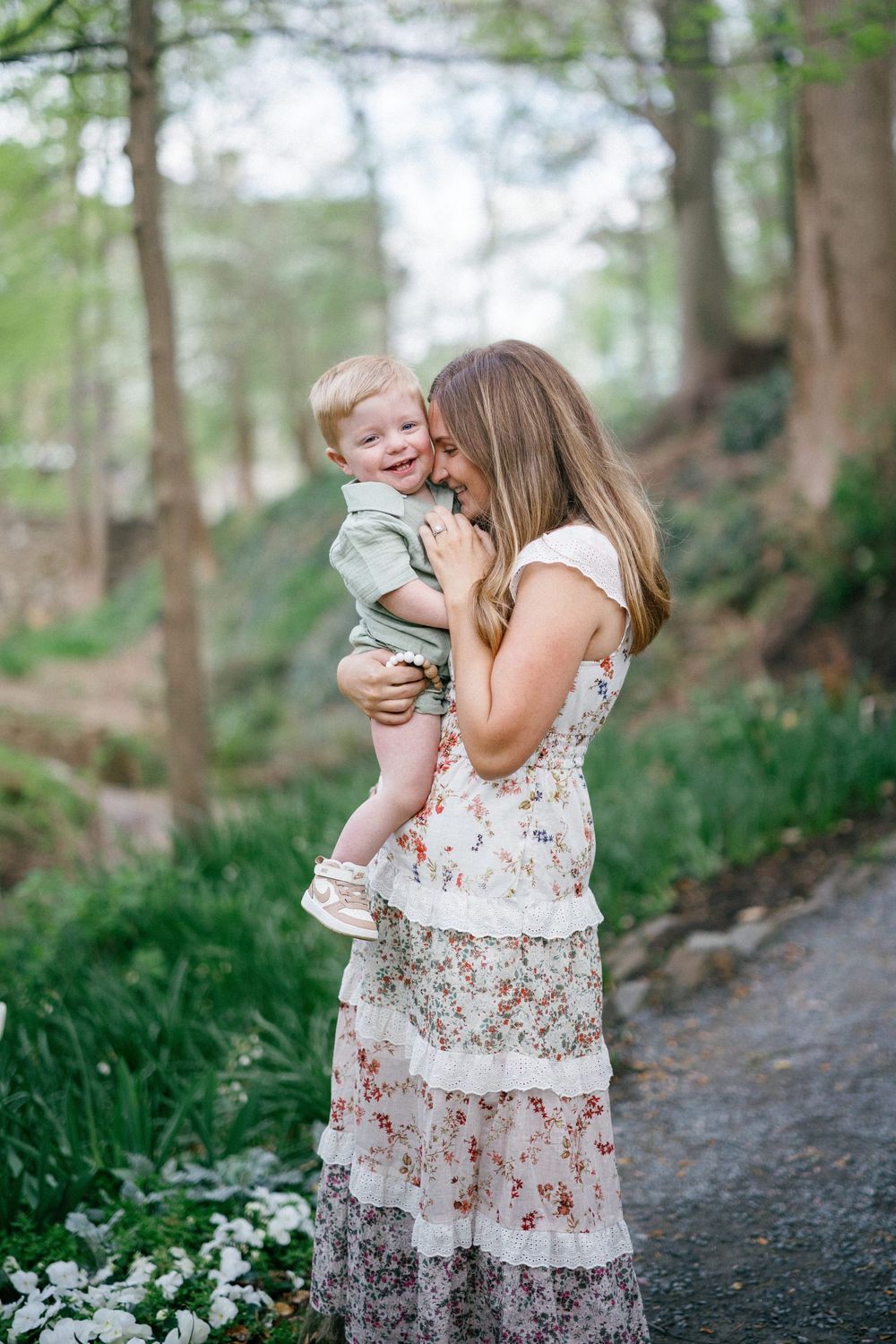 A mother in a floral dress holds her laughing toddler in a lush green garden setting.