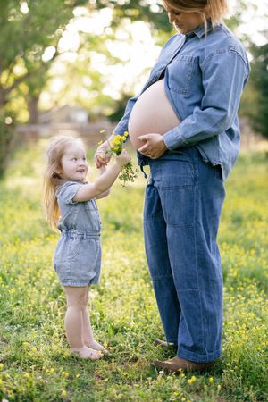 A young child in denim overalls offers wildflowers to a pregnant person wearing denim in a sunny garden setting.