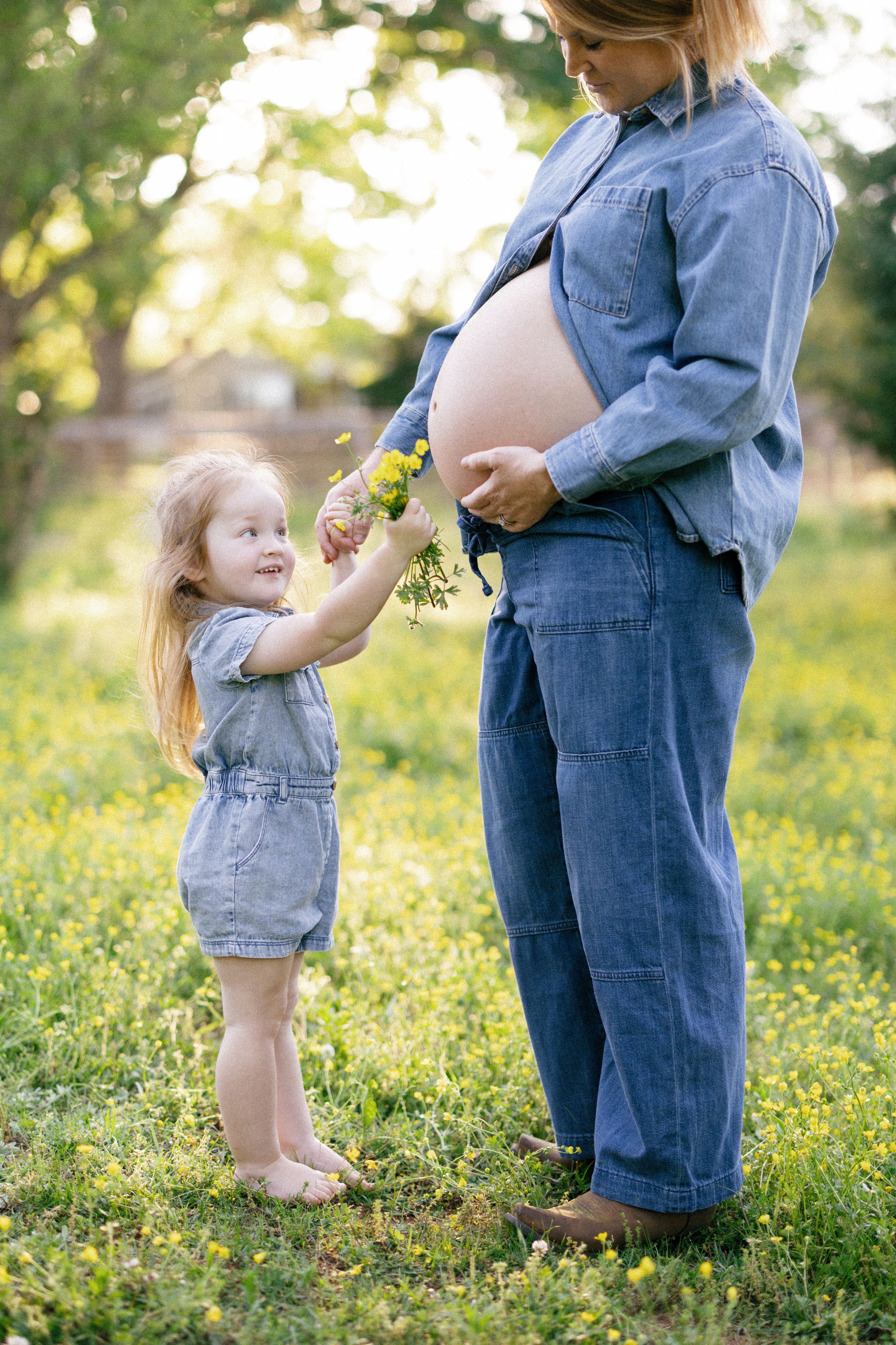 A young child in denim overalls offers wildflowers to a pregnant person wearing denim in a sunny garden setting.