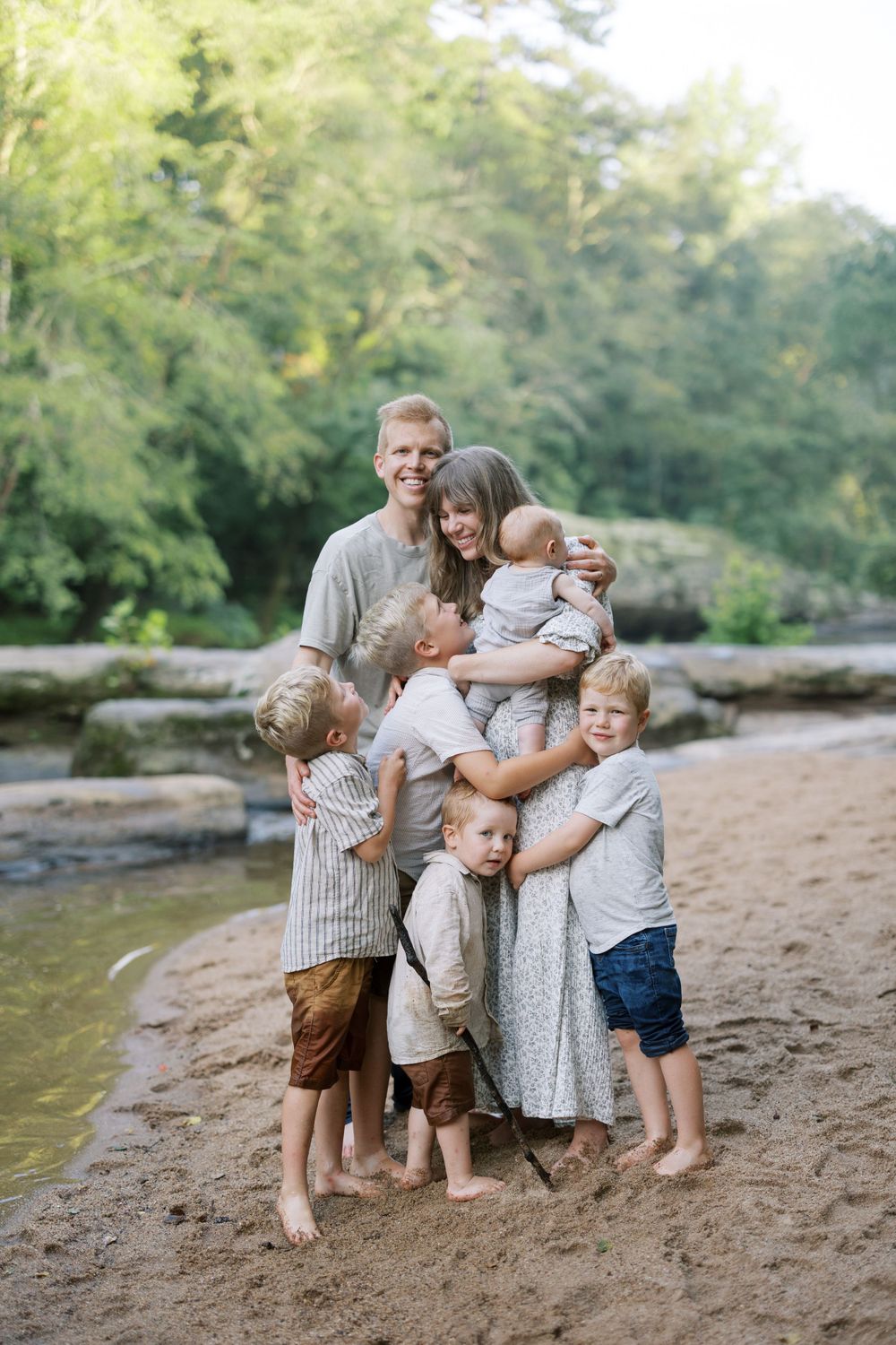 Family group hugging together on sandy riverbank with green forest backdrop.