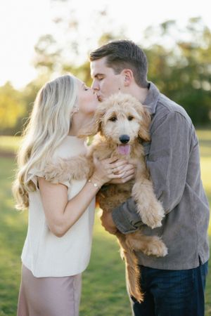 A couple shares a romantic moment while holding their fluffy goldendoodle puppy in a sun-lit outdoor setting.