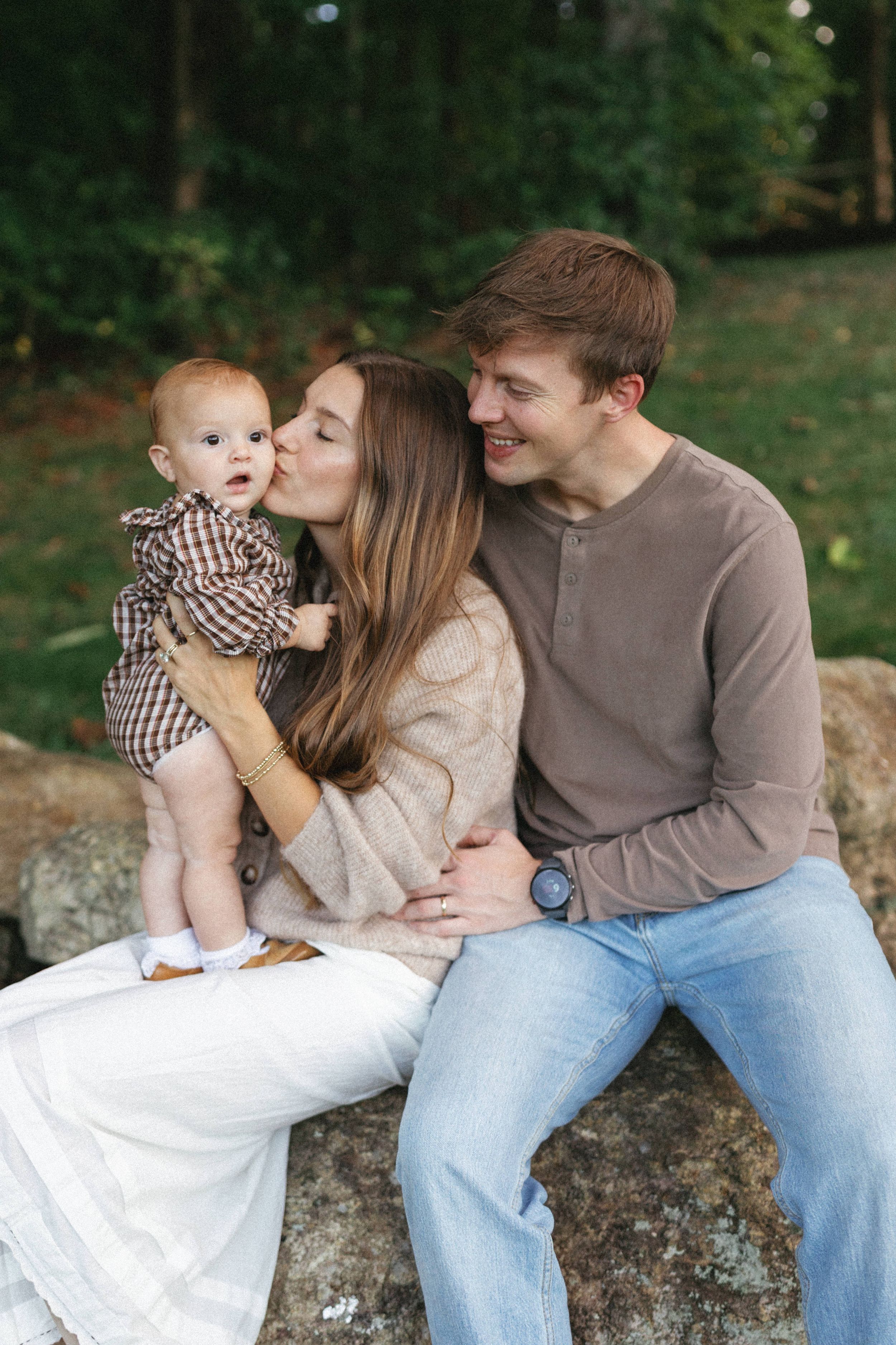 A young family sits on rocks outdoors, mom kissing baby while dad smiles, surrounded by lush green trees.
