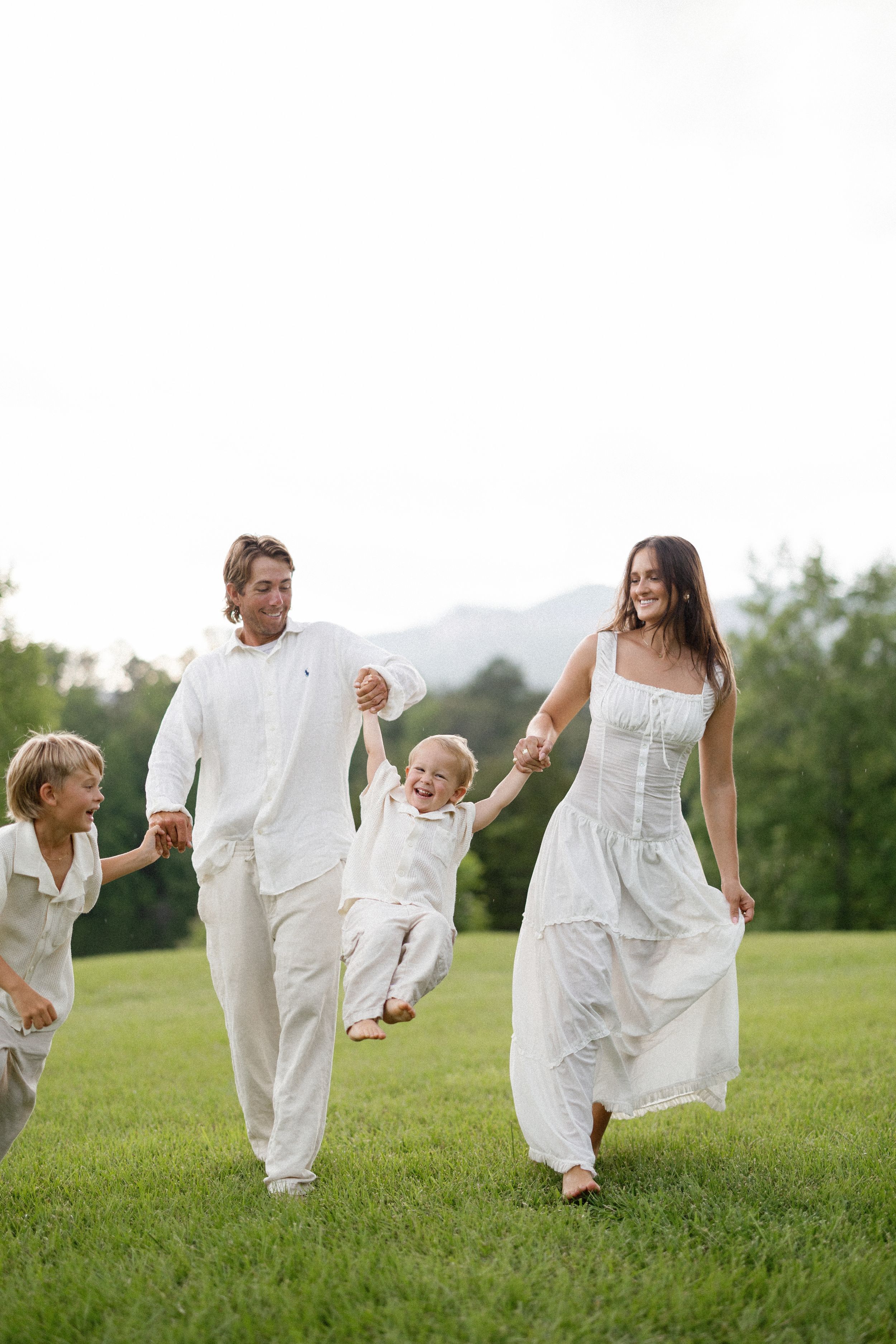 Family wearing white outfits playfully walks together on grassy hill with mountains in background.