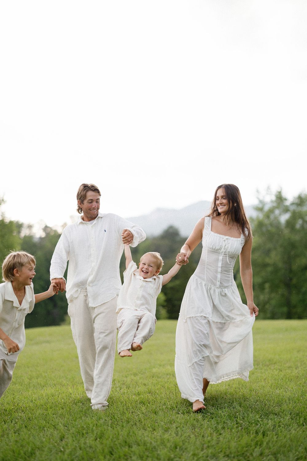 Family wearing white outfits playfully walks together on grassy hill with mountains in background.