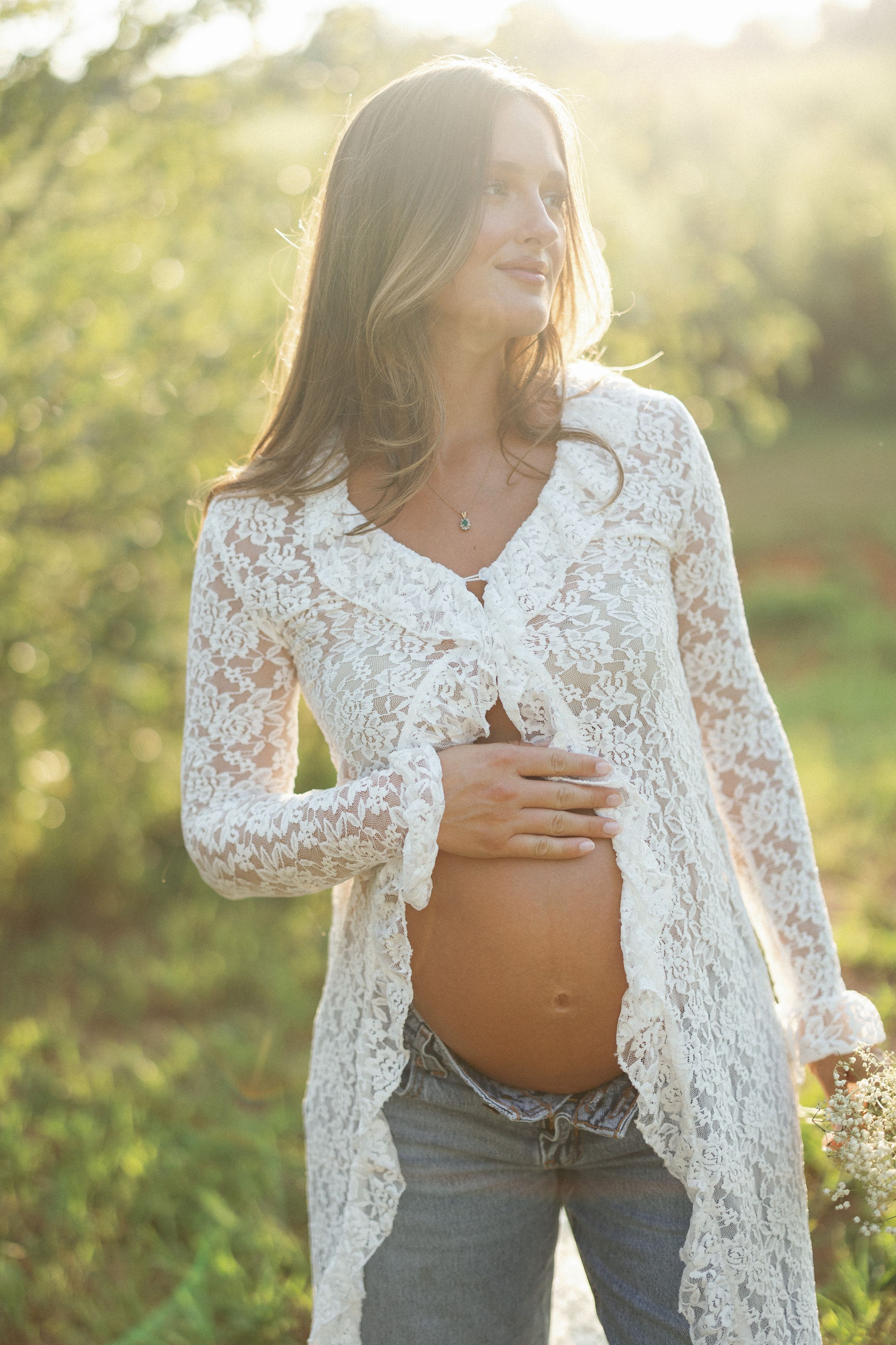 Pregnant woman in white lace cardigan and jeans cradling her baby bump outdoors in golden sunlight.