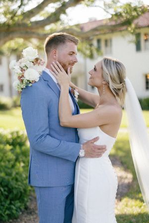 A newlywed couple in blue suit and white dress embrace during an outdoor wedding photoshoot in natural sunlight.