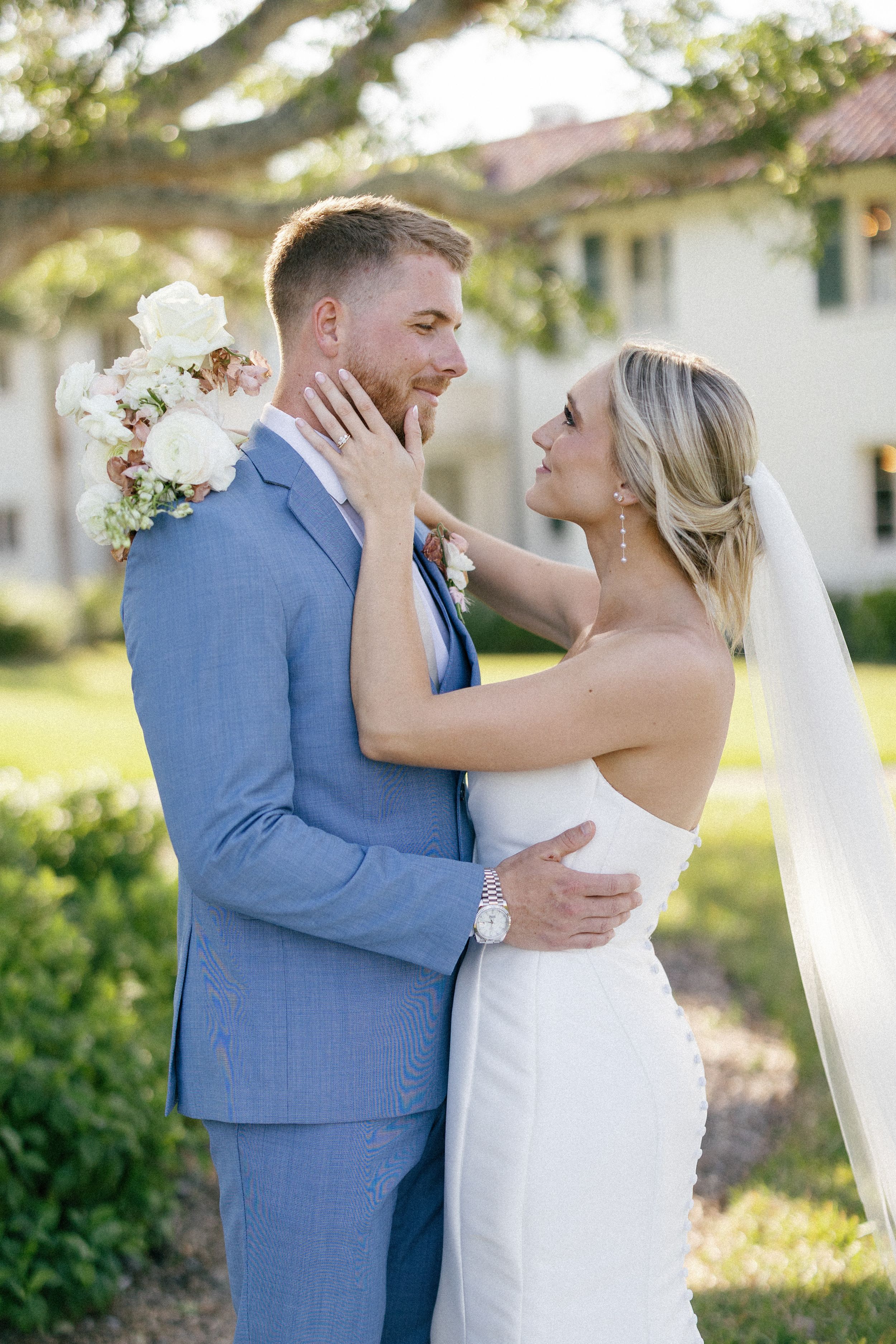 A newlywed couple in blue suit and white dress embrace during an outdoor wedding photoshoot in natural sunlight.