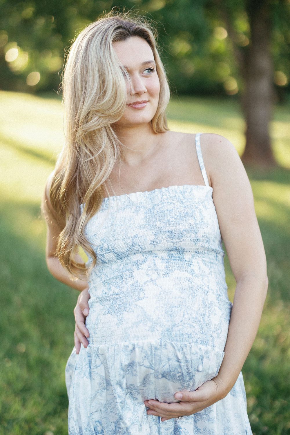 Pregnant woman in floral sundress cradling baby bump outdoors in sunlit park with green trees.