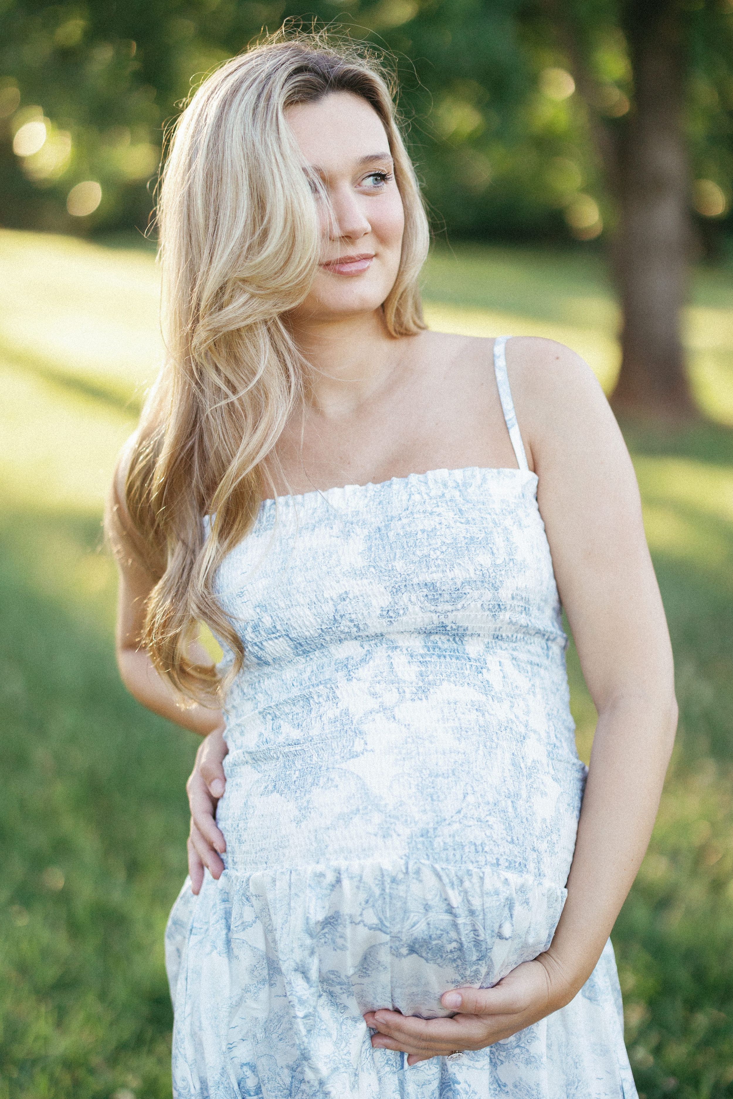 Pregnant woman in floral sundress cradling baby bump outdoors in sunlit park with green trees.