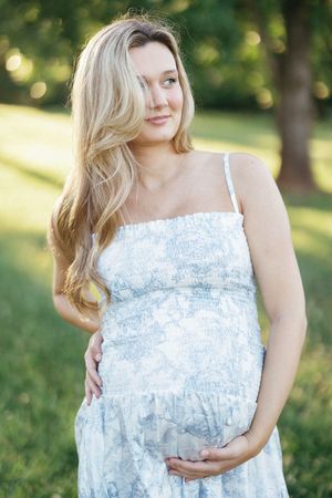 Pregnant woman in floral sundress cradling baby bump outdoors in sunlit park with green trees.
