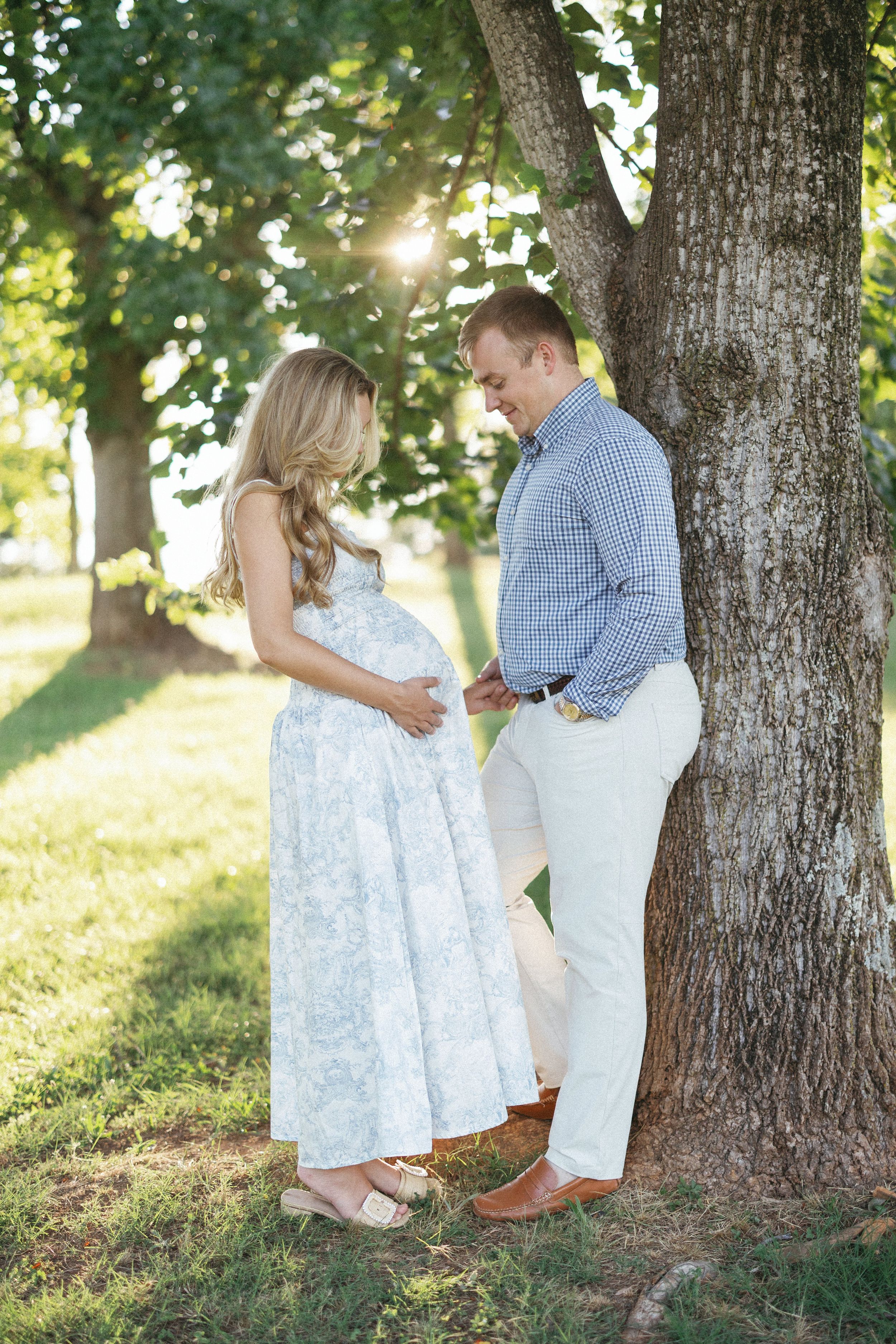 Pregnant couple sharing a tender moment under a sunlit tree, woman in floral dress cradling her baby bump.
