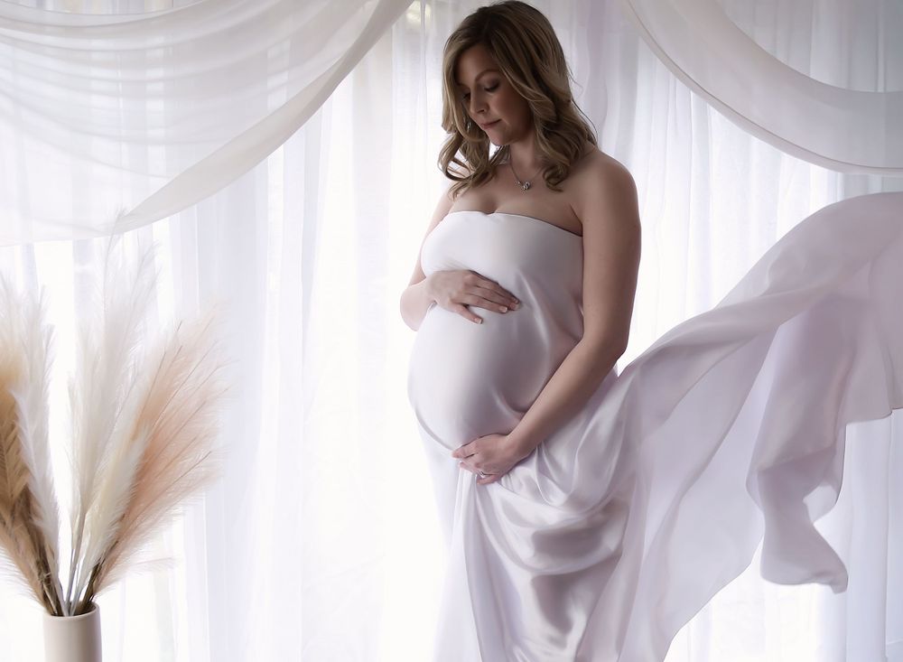 image of mum to be posing in front of a backlit curtain, looking down at her baby bump with hands clasped round.  Fabric falling behind.