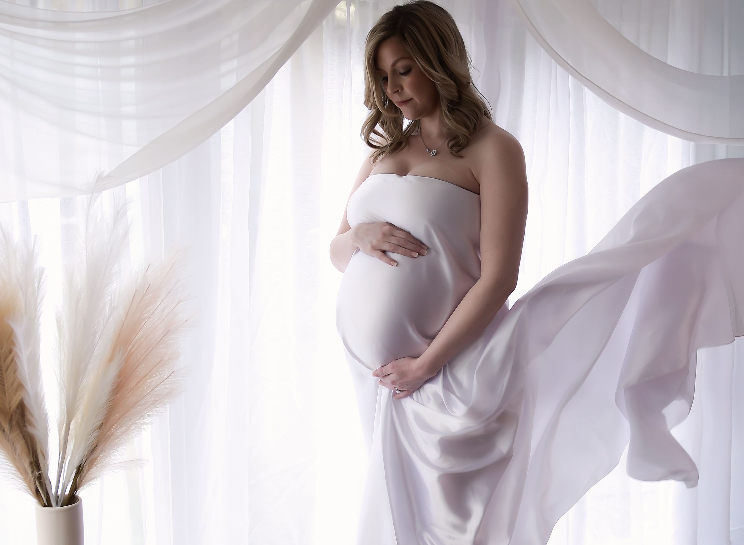 image of mum to be posing in front of a backlit curtain, looking down at her baby bump with hands clasped round.  Fabric falling behind.