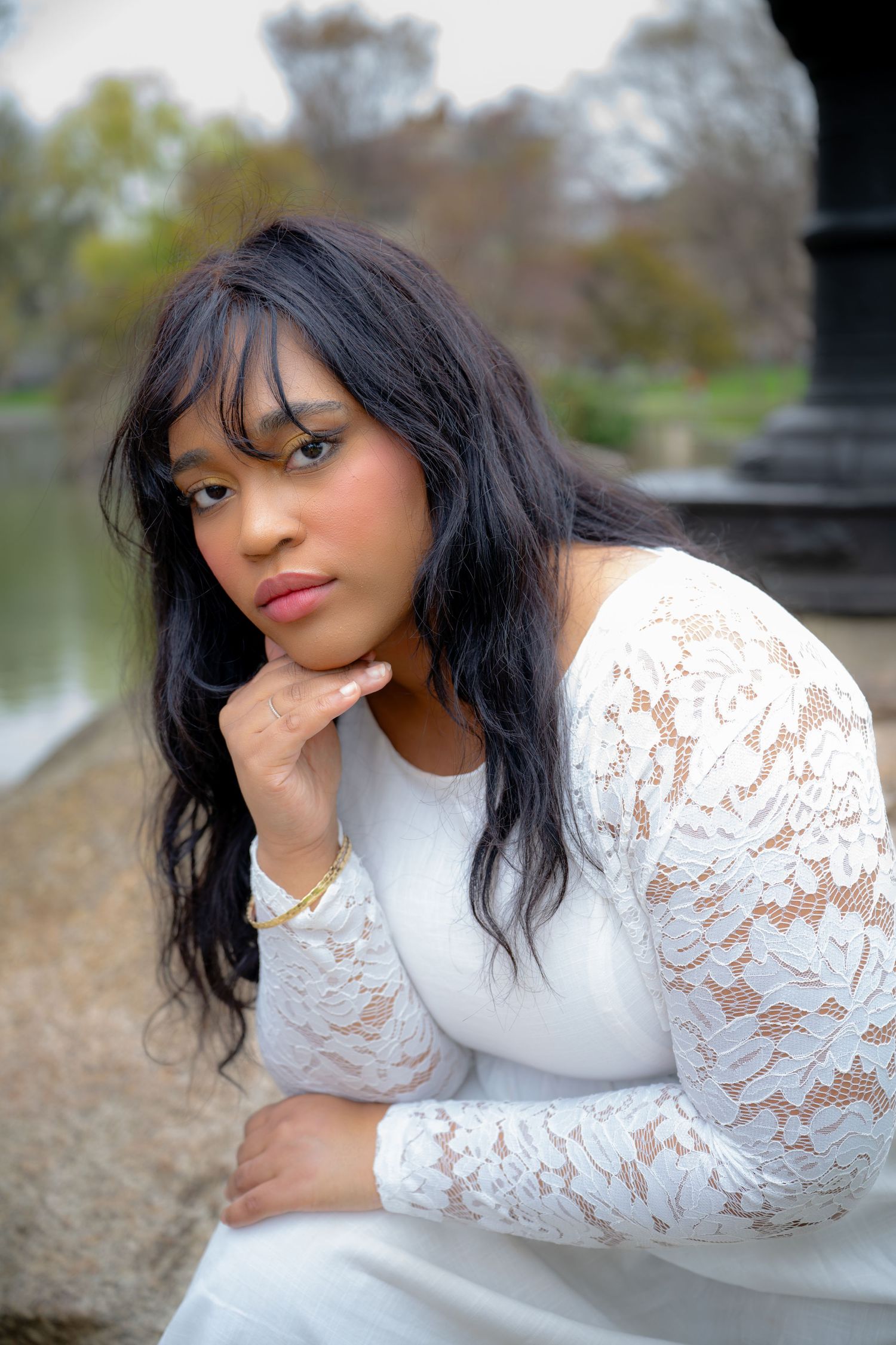 A young woman in a white lace long-sleeve dress sits pensively near a stone column outdoors.