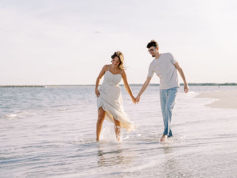 A couple splashing and holding hands while walking through ocean waves on a beach.
